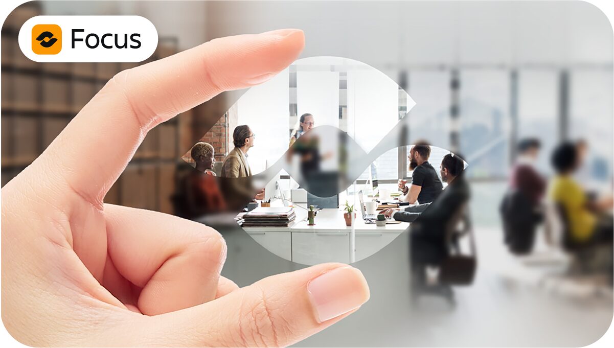 Atlassian Focus Schaubild: Im Hintergrund ist eine unscharfe Szene eines Teams im Meeting, im Vordergrund hält eine Hand das Atlassian Focus Logo wie eine Linse durch die das Team scharf und klar zu sehen ist.